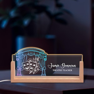 May include: A wooden desk nameplate with a neon blue and purple theater design. The nameplate features the name "Jamie Donovan" and the title "THEATRE TEACHER". The design includes comedy and tragedy masks and a stage curtain.