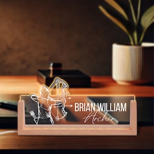 May include: A wooden desk nameplate with a glowing archer design. The nameplate features the name "BRIAN WILLIAM Archer" in white lettering. The archer is depicted in a hooded outfit, drawing a bow. The nameplate is illuminated with a warm light.