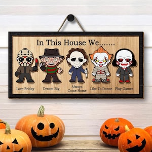 May include: A wooden sign with the text "In This House We..." and five cartoon characters from horror movies. The characters are Jason Voorhees, Freddy Krueger, Michael Myers, Pennywise the Dancing Clown, and Jigsaw. Below each character is a phrase: "Love Friday", "Dream Big", "Always Come Home", "Like To Dance", and "Play Games".