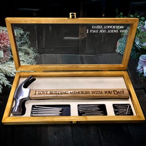 May include: A wooden box with a glass lid, containing a hammer and nails. The hammer handle is engraved with the text "I LOVE BUILDING MEMORIES WITH YOU DAD!". The lid has text in reverse. The box is made of light-coloured wood.