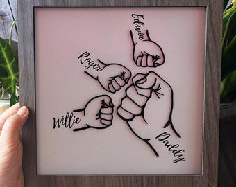 May include: A framed piece of art with a pink background and black silhouettes of five hands in a fist bump. The hands are labeled with the names "Edwin", "Roger", "Willie", "Daddy", and "Daddy".