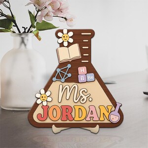 May include: A wooden teacher gift in the shape of a beaker, with the name "Ms. JORDAN" in colourful letters. The design includes a smiling flower, an open book, and science-themed elements.