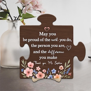 Pode incluir: Peça de quebra-cabeça de madeira marrom com flores e o texto "May you be proud of the work you do, the person you are, and the difference you make. Thank you, Ms. Sophia." Com suporte de madeira.