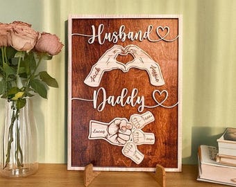 May include: Wooden sign with a dark brown background and a light wood frame. The sign features the words "Husband" and "Daddy" in cursive, along with hand-shaped designs and names. A heart shape is included.