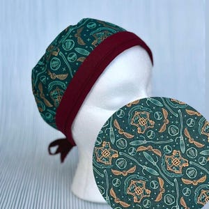 May include: A scrub cap featuring a dark green fabric with a repeating pattern of gold and white crests, winged balls, and broomsticks. The cap has a solid dark maroon band along the bottom edge and ties at the back for an adjustable fit.