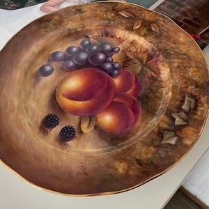 May include: A decorative ceramic plate with a brown and gold background. The plate features a painted design of a bowl of fruit, including peaches and grapes, with blackberries on the rim.