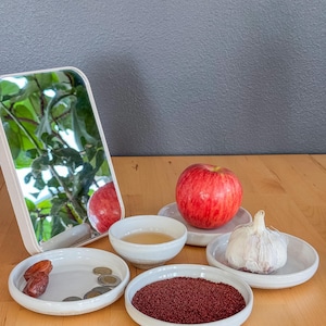 May include: A still life arrangement featuring a rectangular mirror, a red apple, a head of garlic, and small white dishes holding dates, coins, and a reddish-brown spice. A small bowl contains a liquid. The items are on a wooden surface.