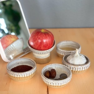 May include: A collection of small, handmade ceramic bowls with a textured, off-white and tan design. The bowls contain various items, including a red apple, dates, coins, and a head of garlic. The items are arranged on a light-colored wooden surface.