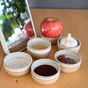 May include: A set of three ceramic mugs with a speckled, matte finish. The mugs are in shades of cream, light gray, and dark gray. Each mug has a simple, minimalist design and a rounded handle.
