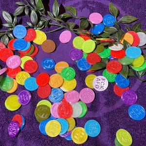 Könnte beinhalten: Eine Ansammlung bunter, runder Spielmarken in verschiedenen Farben, darunter Rot, Blau, Gelb und Grün, auf einer violetten Oberfläche verstreut. Einige Spielmarken tragen den Text "FCK AFD". Grüne Blätterzweige sind um die Spielmarken angeordnet.