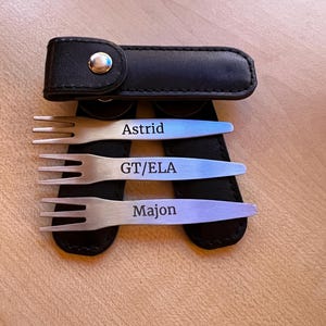 Könnte beinhalten: Drei silberne Gabeln mit schwarzen Ledergriffen. Die Gabeln sind mit den Namen "Astrid", "GT/ELA" und "Majon" graviert.
