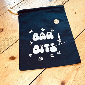Op de afbeelding: Een zwarte trekkoordtas met witte tekst die "Bar Bits" zegt en verschillende kleine witte illustraties bevat, waaronder een kroon, een bloem, een regenboog en een pootafdruk.