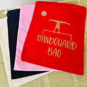 May include: Four drawstring bags stacked on top of each other. The top bag is red with gold text that says "Handguard Bag" and a silhouette of a gymnast on a bar. The next bag is pink, the next is black, and the bottom bag is a natural colour.