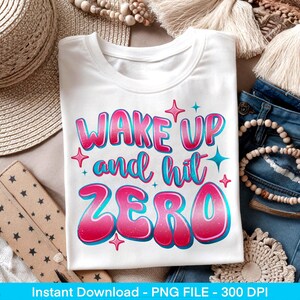 May include: White t-shirt with the text "WAKE UP and hit ZERO" in pink and blue bubble letters. The design includes star accents. The shirt is folded and displayed with denim jeans and accessories.