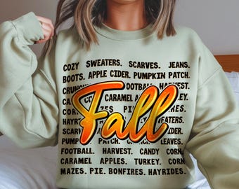 Può includere: Una felpa verde chiaro con la scritta "Fall" in arancione e giallo. Il design include parole a tema autunnale come "zucca", "raccolto" e "mele caramellate".