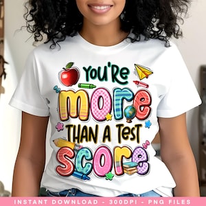 Puede incluir: Camiseta blanca con la frase "You're More Than A Test Score" en letras coloridas y divertidas. El diseño incluye una manzana, un globo terráqueo y útiles escolares, ideal para profesores o estudiantes.