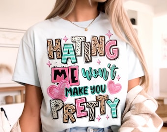 May include: White t-shirt with the text "HATING ME won't MAKE YOU PRETTY" in a colorful, playful design. The text incorporates various patterns, including animal print and stripes, with pink hearts and stars. The shirt is a casual, everyday wear item.