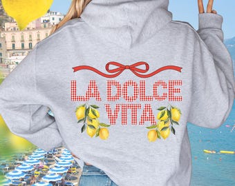 Peut inclure: Un sweat à capuche gris clair avec l'inscription "LA DOLCE VITA" en carreaux rouges. Le motif comprend un ruban rouge et des illustrations de citrons. En arrière-plan, une scène de plage avec des parasols et la mer.