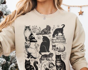 Op de afbeelding: Beige sweatshirt met illustraties van katten in verschillende poses. De katten zijn in zwart-wit, met bloemmotieven. Het ontwerp bevat gedetailleerde tekeningen en silhouetten.