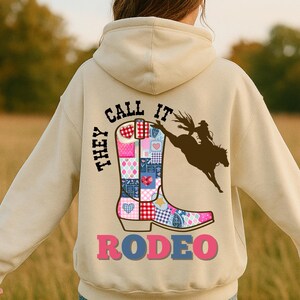 May include: Beige hoodie with a graphic on the back featuring a patchwork cowboy boot, a rodeo rider, and the text "THEY CALL IT RODEO". The boot design includes hearts and various patterns.