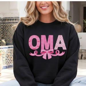 May include: Black sweatshirt with the word "OMA" in pink, featuring a plaid pattern and a bow design. The letters are outlined in a darker pink, with a pink bow at the bottom.