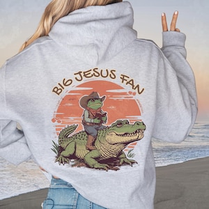 May include: A light gray hoodie with the text "BIG JESUS FAN" in a circular design. The graphic features a frog in a cowboy hat riding an alligator. The frog is holding a book. The background is an orange sunset.