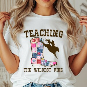 May include: White t-shirt with the text "TEACHING THE WILDEST RIDE". The shirt features a colorful patchwork cowboy boot graphic and a silhouette of a rodeo rider on a bucking horse. A fun and unique design.