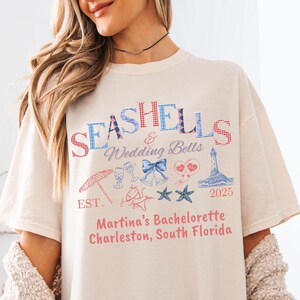 May include: Cream-colored t-shirt with the words "SEASHELLS & Wedding Bells" in blue and red. The shirt features illustrations of a parasol, champagne glasses, bells, hearts, a lighthouse, and starfish. Text at the bottom reads "Martina's Bachelorette Charleston, South Florida."
