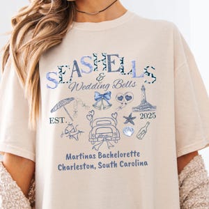 Puede incluir: Camiseta color crema con texto e ilustraciones azules. La camiseta presenta las palabras "Seashells & Wedding Bells" e incluye dibujos de un coche, un faro y otros elementos con temática de playa. También está impreso el texto "Martinas Bachelorette Charleston, South Carolina".