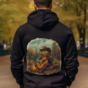 May include: Black hooded sweatshirt featuring a whimsical illustration of a frog cowboy playing a guitar by a campfire. The frog wears a brown hat, red bandana, and smokes a cigarette. The artwork is set against a backdrop of a forest and sky.