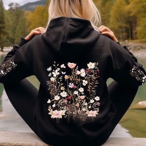 Pu&ograve; includere: Felpa con cappuccio nera con una stampa floreale sul retro. La stampa presenta una variet&agrave; di fiori in tonalit&agrave; rosa, bianco e marrone.