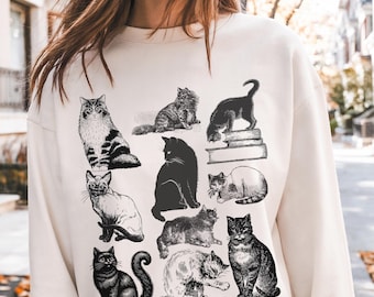 Op de afbeelding: Crèmekleurige sweatshirt met een verzameling zwart-wit kattenillustraties. De katten worden in verschillende poses afgebeeld, waaronder zitten, staan en loungen. Het grafische ontwerp is een tekening in vintage-stijl.