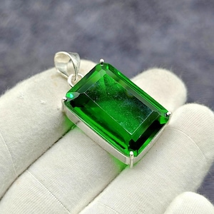 Pode incluir: Pingente retangular de pedra preciosa verde esmeralda com moldura de prata. O pingente tem uma superfície polida e facetada que reflete a luz. O pingente é preso por uma argola de prata.