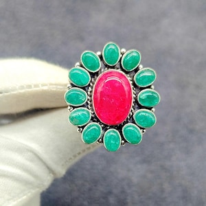 May include: A silver ring featuring a large oval, bright red gemstone at the center, surrounded by a halo of smaller, oval-shaped, green gemstones. The ring has a detailed, textured silver band.