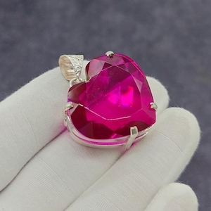 May include: A heart-shaped pendant featuring a large, faceted, magenta gemstone. The gemstone is set in a silver-coloured metal frame with a decorative bail. The pendant is held by a gloved hand against a dark grey background.