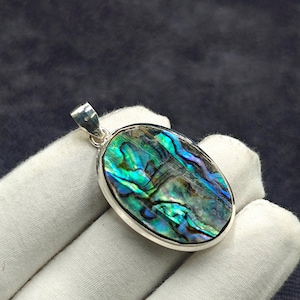 Puede incluir: Colgante ovalado con incrustación de concha de abulón iridiscente, mostrando azules, verdes y púrpuras. La concha está engastada en un marco plateado pulido, con una argolla plateada para una cadena de collar.