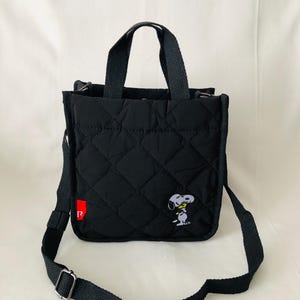 May include: A black quilted tote bag with two top handles and an adjustable shoulder strap. The bag features a diamond-stitched pattern, a small red tag with the letter R on the side, and an embroidered graphic of Snoopy on the lower right.