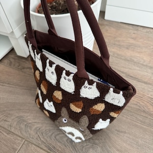 May include: A brown tote bag featuring a textured, fuzzy surface with a repeating pattern of small white creatures and brown acorns. The bag has two dark brown fabric handles and a larger, grey creature graphic at the bottom front section.