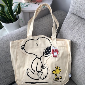 May include: A cream-coloured canvas tote bag featuring a large black line-art illustration of Snoopy and a smaller yellow illustration of Woodstock. A small tag shaped like a red doghouse is attached to the handle, and a small white brand tag is on the side.