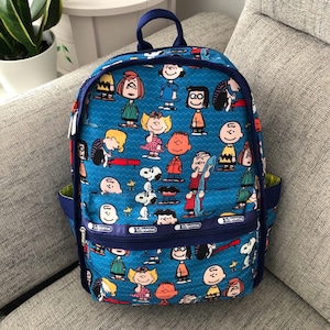 May include: A blue backpack featuring a repeating pattern of Peanuts characters including Snoopy, Charlie Brown, and Lucy. The bag has a dark blue zip, a front pocket with the LeSportsac logo, a top handle, and a subtle wavy line background texture.