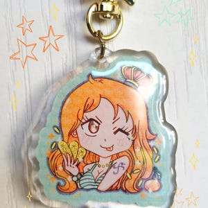 Peut inclure: Un porte-clés en acrylique transparent avec une illustration de dessin animé d'un personnage aux cheveux orange, faisant un clin d'œil et tenant des pièces d'or. Le porte-clés a un fermoir doré et un bord bleu clair et orange avec des étoiles.