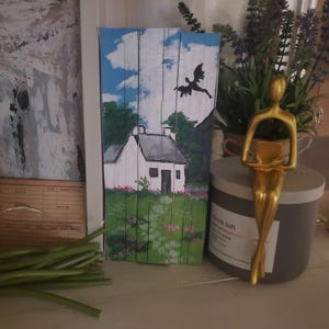 Può includere: Opera d'arte in legno raffigurante un cottage bianco con tetto grigio, sullo sfondo di un cielo blu con nuvole. Una silhouette nera di un drago vola sopra il cottage. L'opera è su una tavola di legno.
