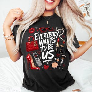 Peut inclure: T-shirt noir avec l'inscription "EVERYBODY WANTS TO BE US" et des illustrations de rouge à lèvres rouge, d'un sac à main, de talons hauts, de lunettes de soleil et d'autres articles de beauté. Le design comprend des cœurs et un téléphone portable.