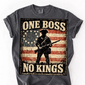 Peut inclure: T-shirt gris foncé avec un motif vieilli d'un drapeau américain et une silhouette de soldat tenant un fusil. Le texte "ONE BOSS NO KINGS" est imprimé au-dessus et en dessous du motif du drapeau. Le t-shirt a des manches retroussées.