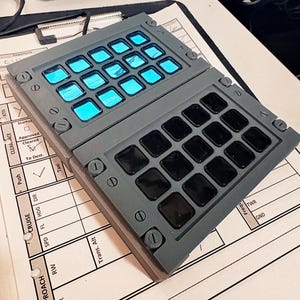 May include: A grey, rectangular, two-piece keypad with a series of black and blue buttons. The top piece has blue buttons with white text, including "15:56". The bottom piece has black buttons.