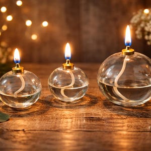 Peut inclure: Trois lampes à huile sphériques en verre de tailles différentes, chacune dotée d'un support de mèche en métal doré et d'une flamme allumée. Les récipients en verre transparent contiennent du combustible liquide et une mèche blanche.