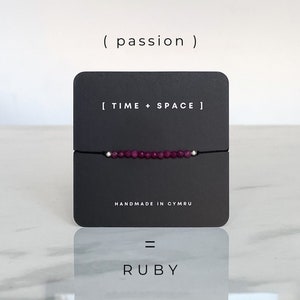 May include: A black card with the text "[ TIME + SPACE ]" and "HANDMADE IN CYMRU" on it. A red gemstone bracelet is displayed on the card. The text "( passion )" is above the card and the text "RUBY" is below the card.