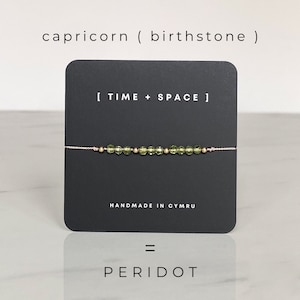 Puede incluir: Un brazalete de piedra de peridoto verde en una cuerda rosa con cuentas doradas. El brazalete está sobre una tarjeta negra con el texto "[ TIME + SPACE ]" y "HANDMADE IN CYMRU". La tarjeta también tiene el texto "capricorn ( birthstone )" y "PERIDOT".