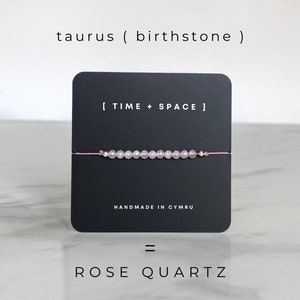 May include: A pink rose quartz bracelet with a black card that says "[TIME + SPACE]" and "HANDMADE IN CYMRU". The card is on a white marble surface. The text "taurus (birthstone)" and "= ROSE QUARTZ" are also visible.