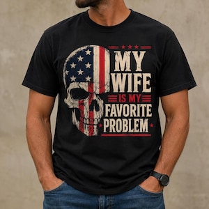 May include: A black short-sleeved t-shirt featuring a graphic of a human skull with a United States flag pattern. The text printed in white and red reads, MY WIFE IS MY FAVORITE PROBLEM, with three stars above and red horizontal lines as accents.